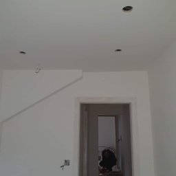 Prywatny wykonawca - Wnętrze w trakcie prac wykończeniowych: białe ściany z widocznymi przewodami elektrycznymi, sufit z otworami na oświetlenie, widoczna osoba pracująca w głębi pomieszczenia przez futrynę drzwiową.