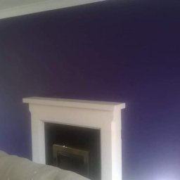 Prywatny wykonawca - Ściana pomalowana na intensywny fioletowy kolor, z białym kominkiem i fragmentem sofy w dolnym rogu, obok widoczne białe drzwi.