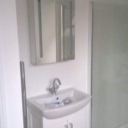 Prywatny wykonawca - Biała umywalka z szafką i lustrem w łazience, widoczna kabina prysznicowa, karton na podłodze sugeruje etap prac wykończeniowych.