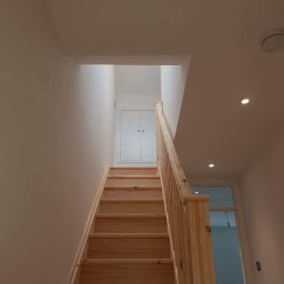 Prywatny wykonawca - Jasne drewniane schody z prostą balustradą w nowo wykończonym wnętrzu, białe ściany i sufit z widocznymi punktowymi lampami.