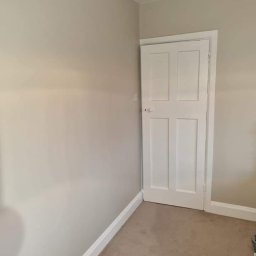 Prywatny wykonawca - Świeżo pomalowane wnętrze pokoju z beżowymi ścianami, białymi listwami przypodłogowymi i białymi drzwiami z czterema panelami, widoczne ślady po malowaniu na dywanie.