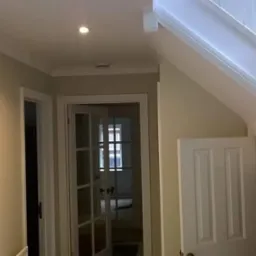 Pomieszczenie po odświeżeniu: ściany pomalowane na jasny beż, białe drzwi wewnętrzne z szybami, białe schody z drewnianą balustradą, sufit z punktowym oświetleniem.