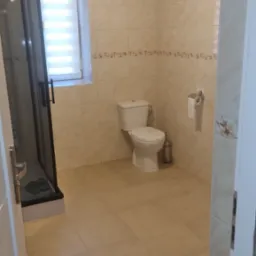 Jasna łazienka z beżowymi płytkami, kabiną prysznicową z czarną ramą, toaletą i oknem z żaluzjami.