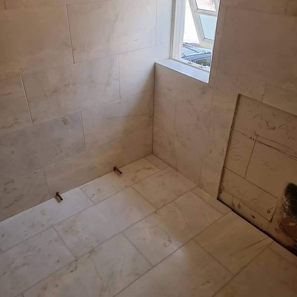 Fragment łazienki w trakcie układania marmurowych płytek, widoczne odstające elementy montażowe i notatki na ścianie.