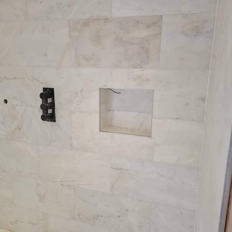 Ściana wyłożona marmurowymi płytkami w łazience, z wnęką w ścianie wykończoną metalową ramką i czarnym, potrójnym zaworem termostatycznym.