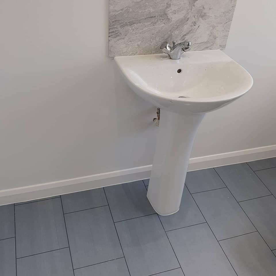 Biała umywalka na postumencie z chromowaną baterią, zamontowana na tle szarej płytki imitującej marmur i szarych podłogowych płytek prostokątnych.