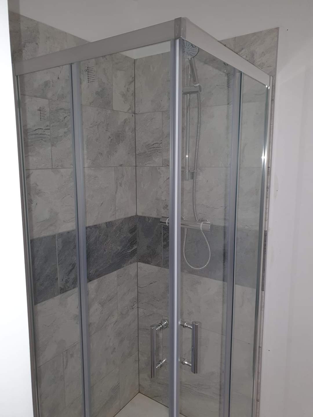 Szklana kabina prysznicowa z chromowanymi uchwytami, zamontowana w narożniku wyłożonym szarymi płytkami imitującymi kamień, z poziomym pasem ciemniejszych płytek.
