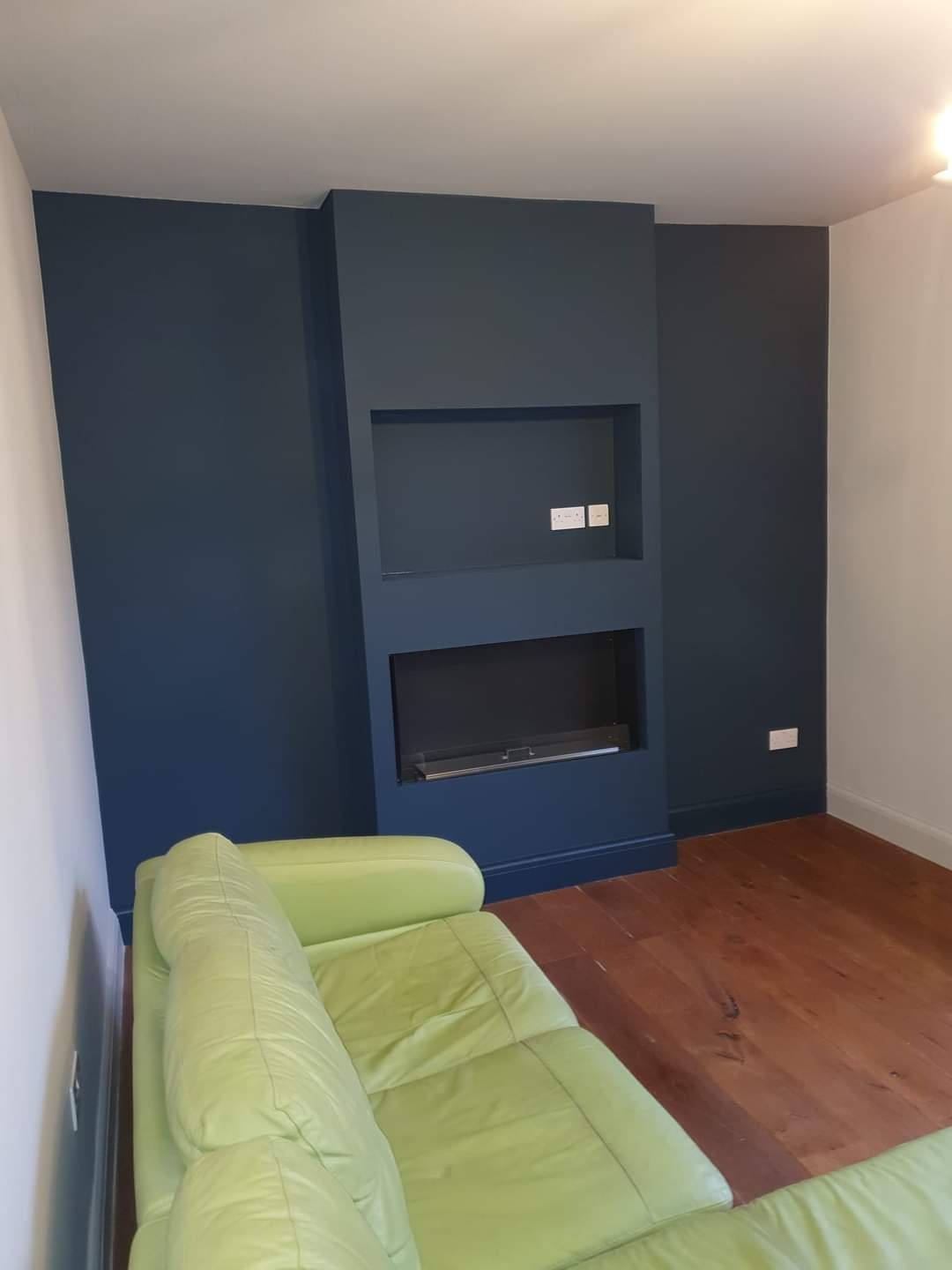 Pomieszczenie po malowaniu ścian na ciemny granat z wnęką na telewizor i kominek elektryczny, drewniana podłoga i jasna kanapa.