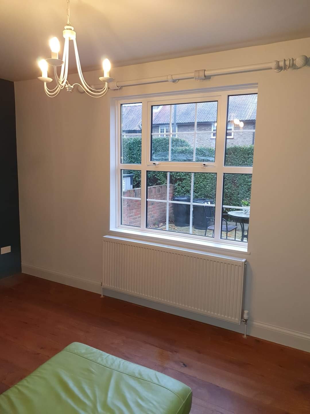 Jasne wnętrze pokoju po remoncie, z drewnianą podłogą, białymi ścianami, oknem z widokiem na ogród i grzejnikiem pod oknem, oświetlone ozdobnym żyrandolem.