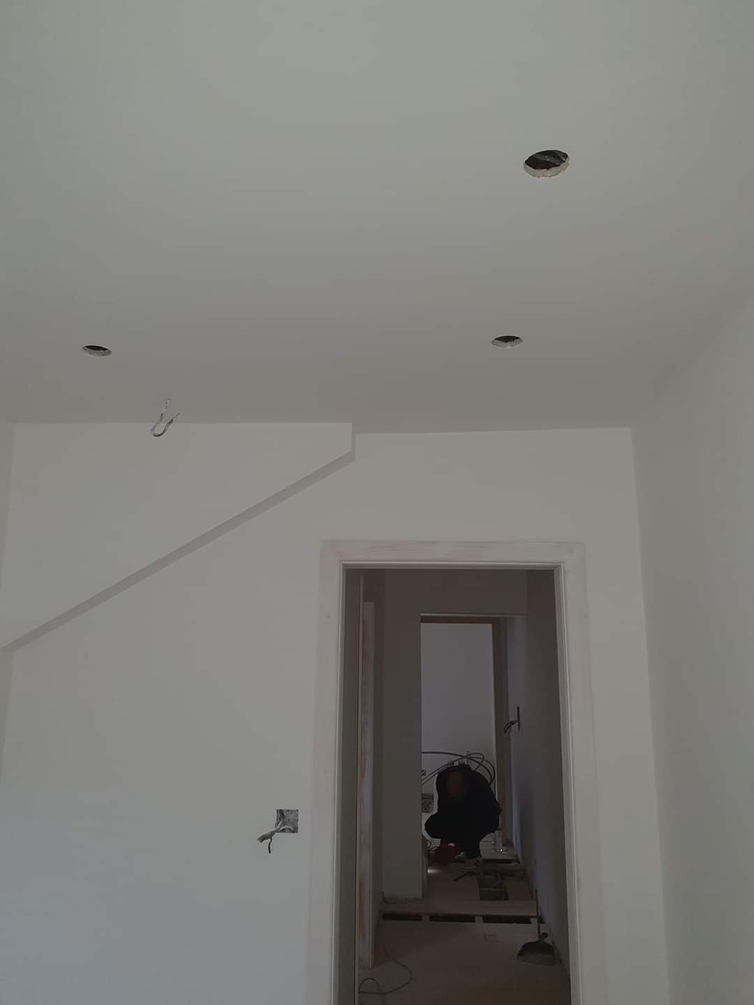 Wnętrze w trakcie prac wykończeniowych: białe ściany z widocznymi przewodami elektrycznymi, sufit z otworami na oświetlenie, widoczna osoba pracująca w głębi pomieszczenia przez futrynę drzwiową.