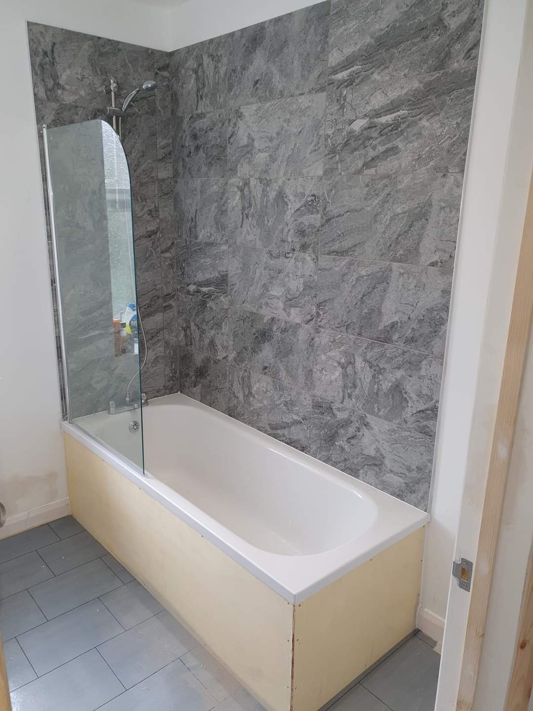 Wanna z zamontowanym parawanem nawannowym, obudowana płytami OSB, szare płytki imitujące marmur na ścianie, szara podłoga z płytek, widoczny fragment niedokończonej futryny drzwi.