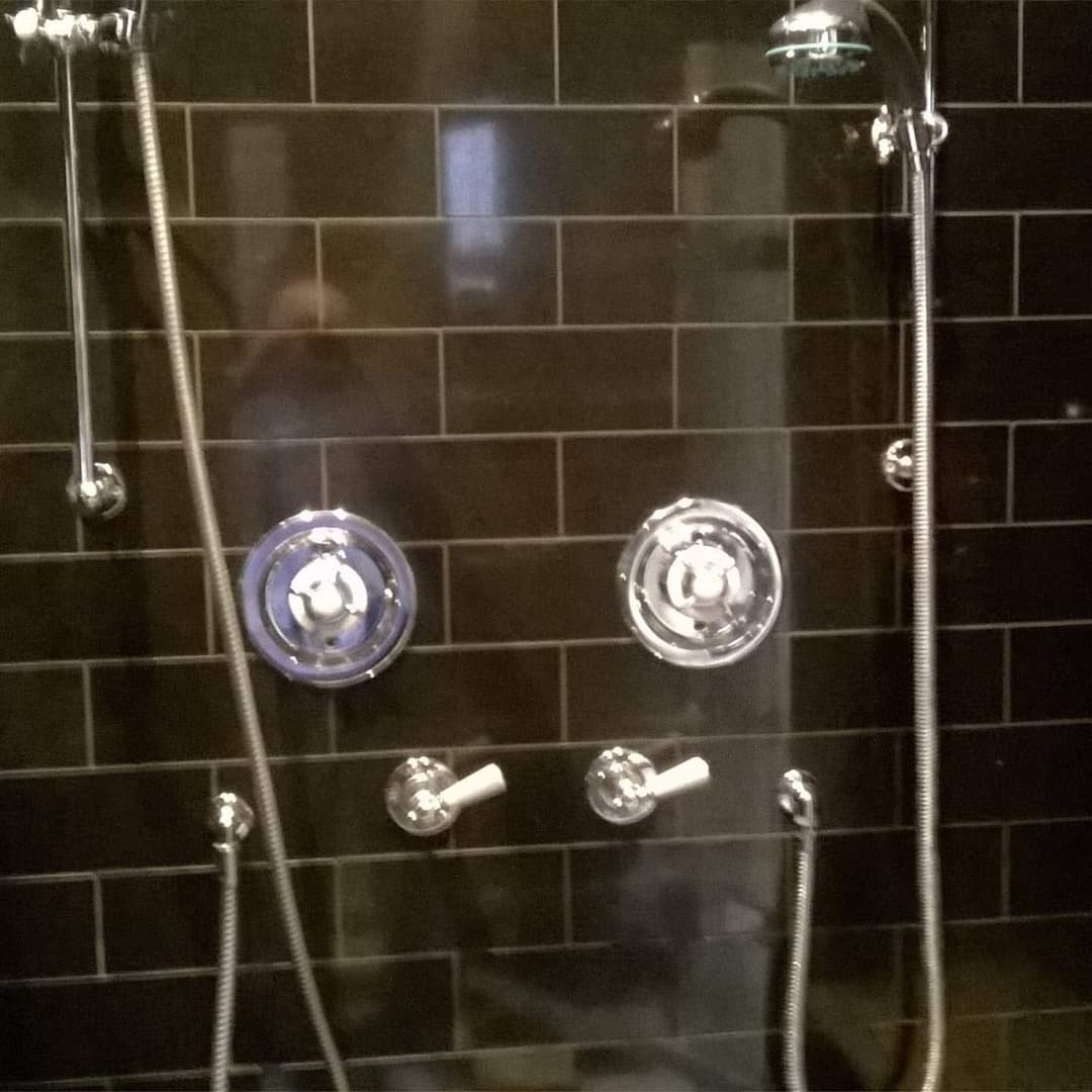 Ciemne, błyszczące płytki w kabinie prysznicowej z chromowaną armaturą i deszczownicą. Widoczne dwa pokrętła, jedno z niebieskim akcentem.