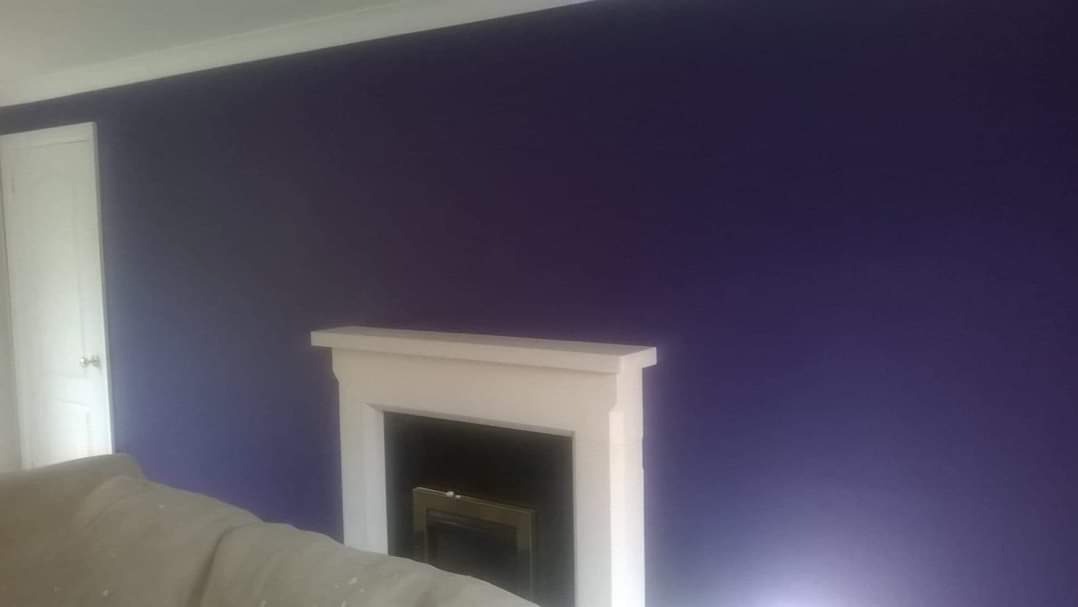 Ściana pomalowana na intensywny fioletowy kolor, z białym kominkiem i fragmentem sofy w dolnym rogu, obok widoczne białe drzwi.
