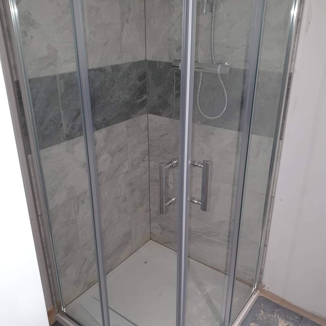 Szklana kabina prysznicowa narożna z chromowanymi uchwytami, wyłożona płytkami w odcieniach szarości, z widoczną słuchawką prysznicową.