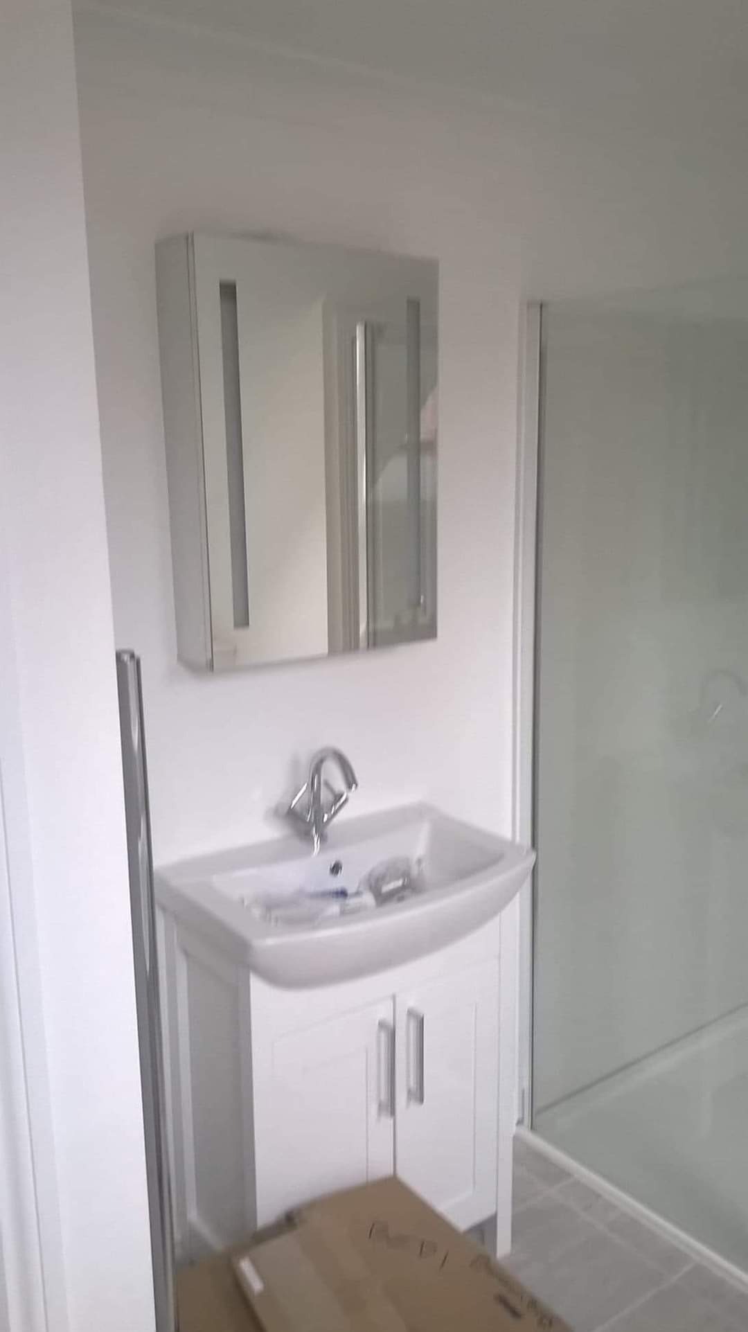 Biała umywalka z szafką i lustrem w łazience, widoczna kabina prysznicowa, karton na podłodze sugeruje etap prac wykończeniowych.