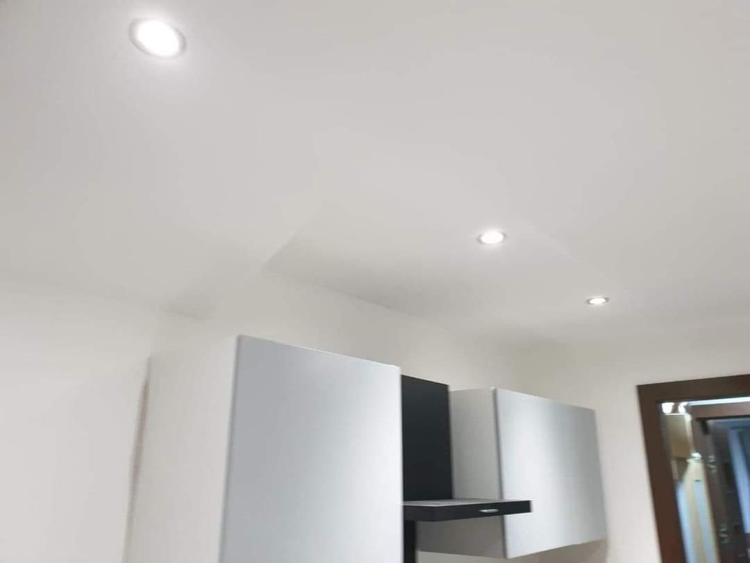 Nowoczesna kuchnia z białymi szafkami górnymi i czarnymi akcentami, oświetlona trzema punktowymi lampami sufitowymi, widok z dołu.