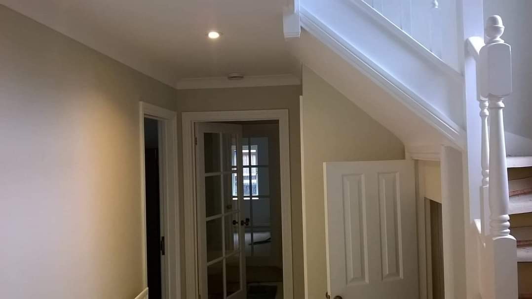 Korytarz po malowaniu w odcieniach beżu i bieli z widocznymi schodami, drzwiami i oświetleniem punktowym.