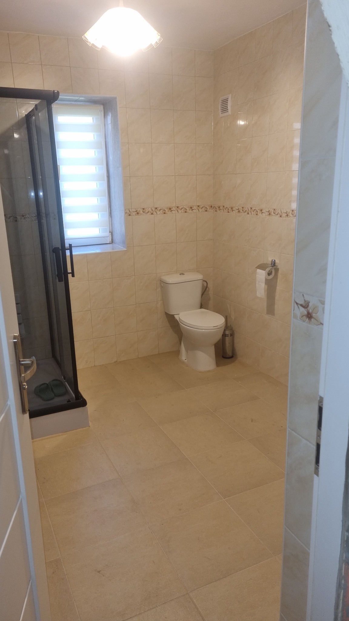 Jasna łazienka z beżowymi płytkami, kabiną prysznicową z czarną ramą, toaletą i oknem z żaluzjami.
