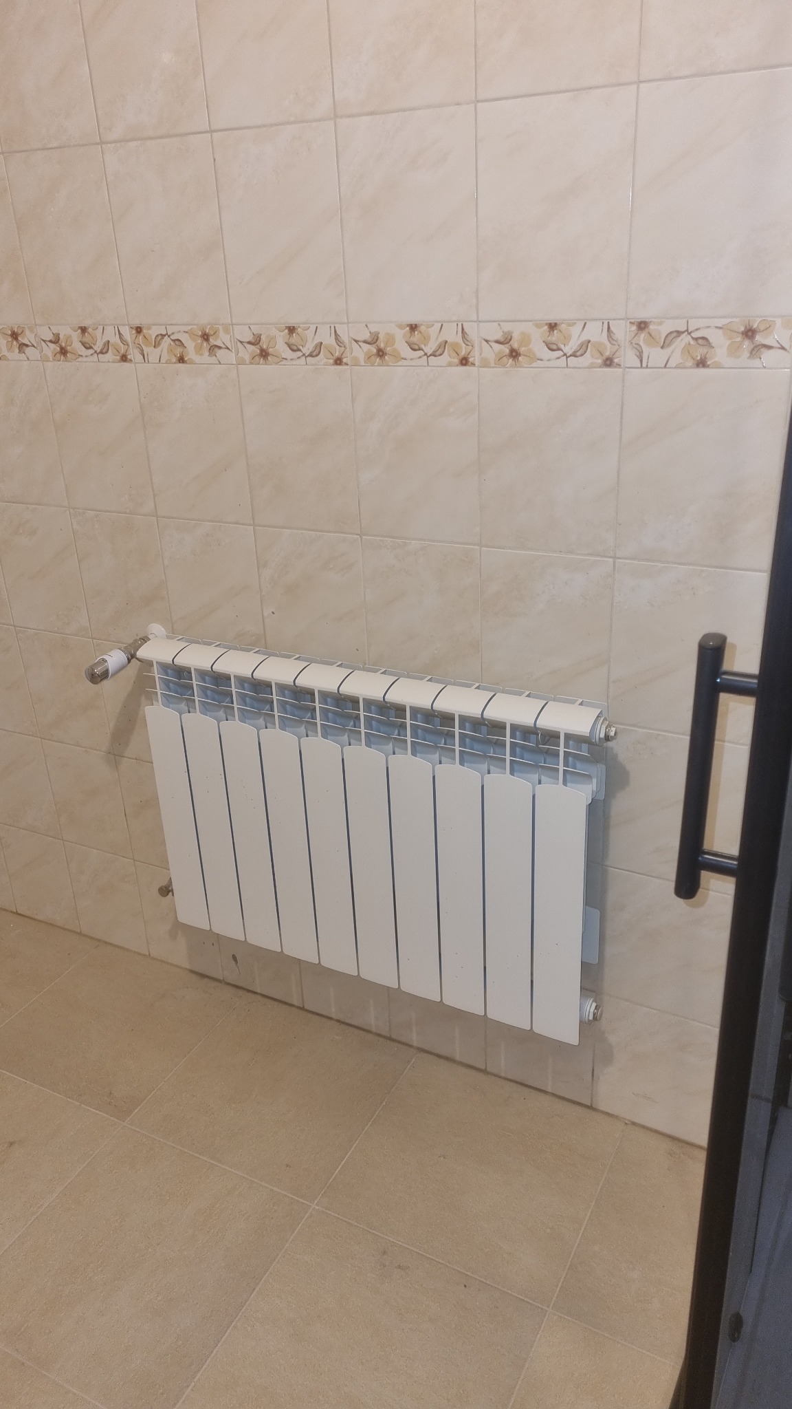 Biały grzejnik panelowy zamontowany na ścianie w łazience wyłożonej beżowymi płytkami z dekoracyjnym pasem kwiatowym. Widoczny zawór termostatyczny i fragment czarnej ramy drzwi.