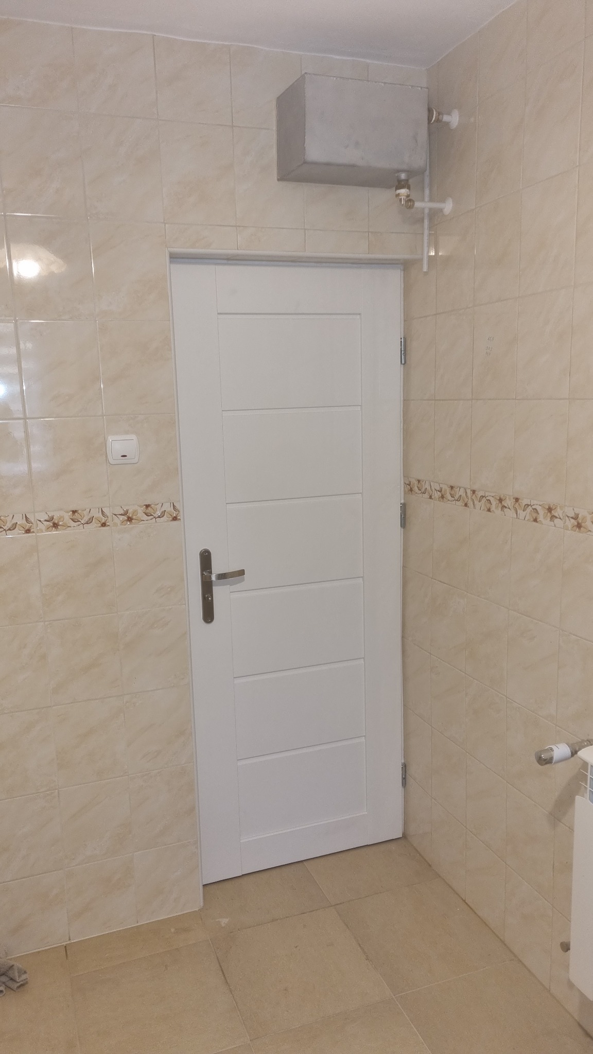 Wnętrze łazienki z beżowymi płytkami, białą, nowoczesną, wewnętrzną stolarką drzwiową, grzejnikiem oraz szarą, metalową obudową instalacji nad drzwiami.