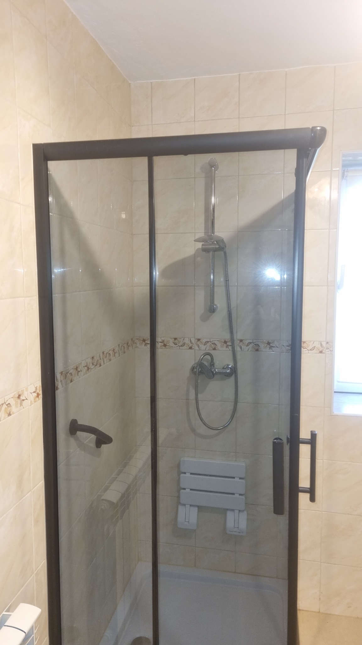 Kabina prysznicowa z brązowymi ramami i siedziskiem, wyłożona beżowymi płytkami z dekoracyjnym pasem, widoczne elementy armatury.