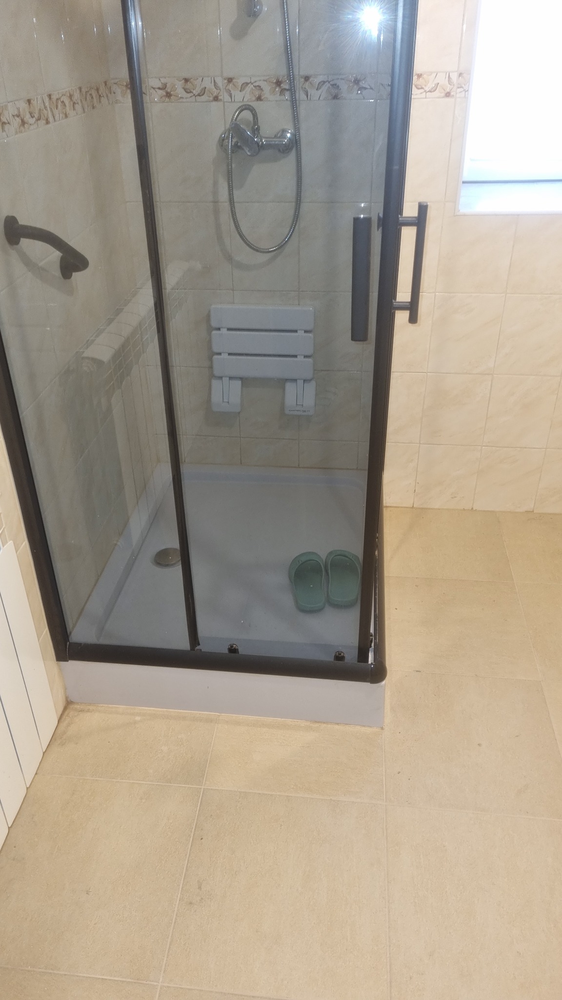 Kabina prysznicowa z siedziskiem i parą zielonych klapek wewnątrz, wykończona beżowymi płytkami z delikatnym kwiatowym wzorem przy suficie, widoczny uchwyt dla niepełnosprawnych.