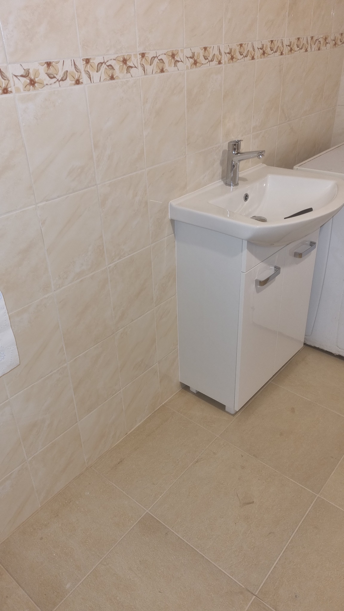 Biała szafka pod umywalką z chromowaną baterią i beżowymi płytkami ściennymi z motywem kwiatowym w łazience.