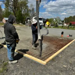 Dwóch pracowników w trakcie wylewania betonu na siatkę zbrojeniową w drewnianej szalunkowej ramie; w tle betoniarka i zabudowa mieszkalna.