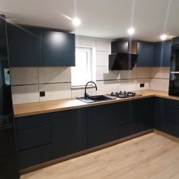 CONSTRUO - Nowoczesna kuchnia z ciemnymi, matowymi szafkami, drewnianym blatem i czarnymi akcentami: zlew, bateria, płyta grzewcza i okap. Duża, czarna lodówka po lewej stronie.