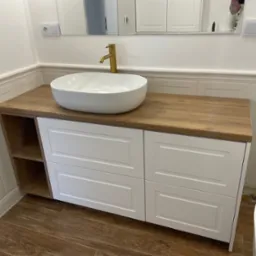 Biała umywalka nablatowa z złotą baterią na drewnianym blacie z białymi szafkami z szufladami i półkami, w tle białe płytki i lustro.