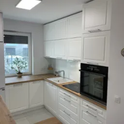 Nowoczesna, jasna kuchnia z białymi frontami szafek, drewnianym blatem i widokiem na zimowy krajobraz za oknem. Na blacie stoi doniczka z małym drzewkiem.