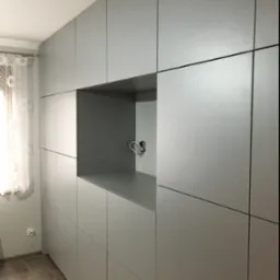 Szara zabudowa meblowa od podłogi do sufitu z otwartą wnęką na sprzęt RTV i wystającymi kablami, widoczna z boku przy ścianie z oknem z firanką.