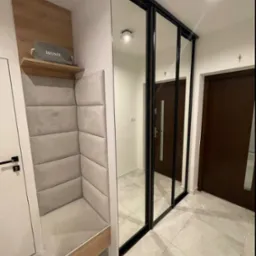 Nowoczesny przedpok&oacute;j z zabudową siedziska z tapicerowanymi panelami, drewnianą p&oacute;łką i lustrzaną szafą przesuwną w czarnej ramie.