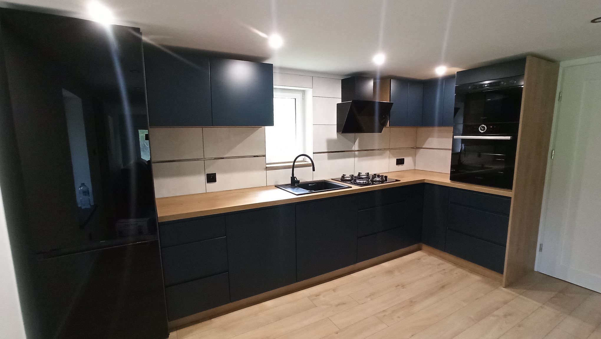 Nowoczesna kuchnia z ciemnymi, matowymi szafkami, drewnianym blatem i czarnymi akcentami: zlew, bateria, płyta grzewcza i okap. Duża, czarna lodówka po lewej stronie.