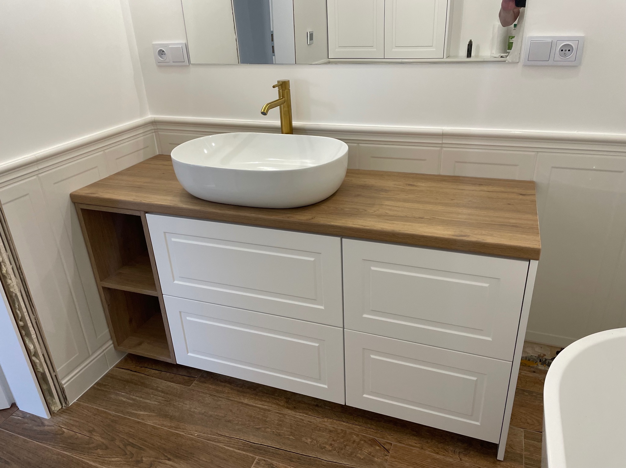 Biała umywalka nablatowa z złotą baterią na drewnianym blacie z białymi szafkami z szufladami i półkami, w tle białe płytki i lustro.