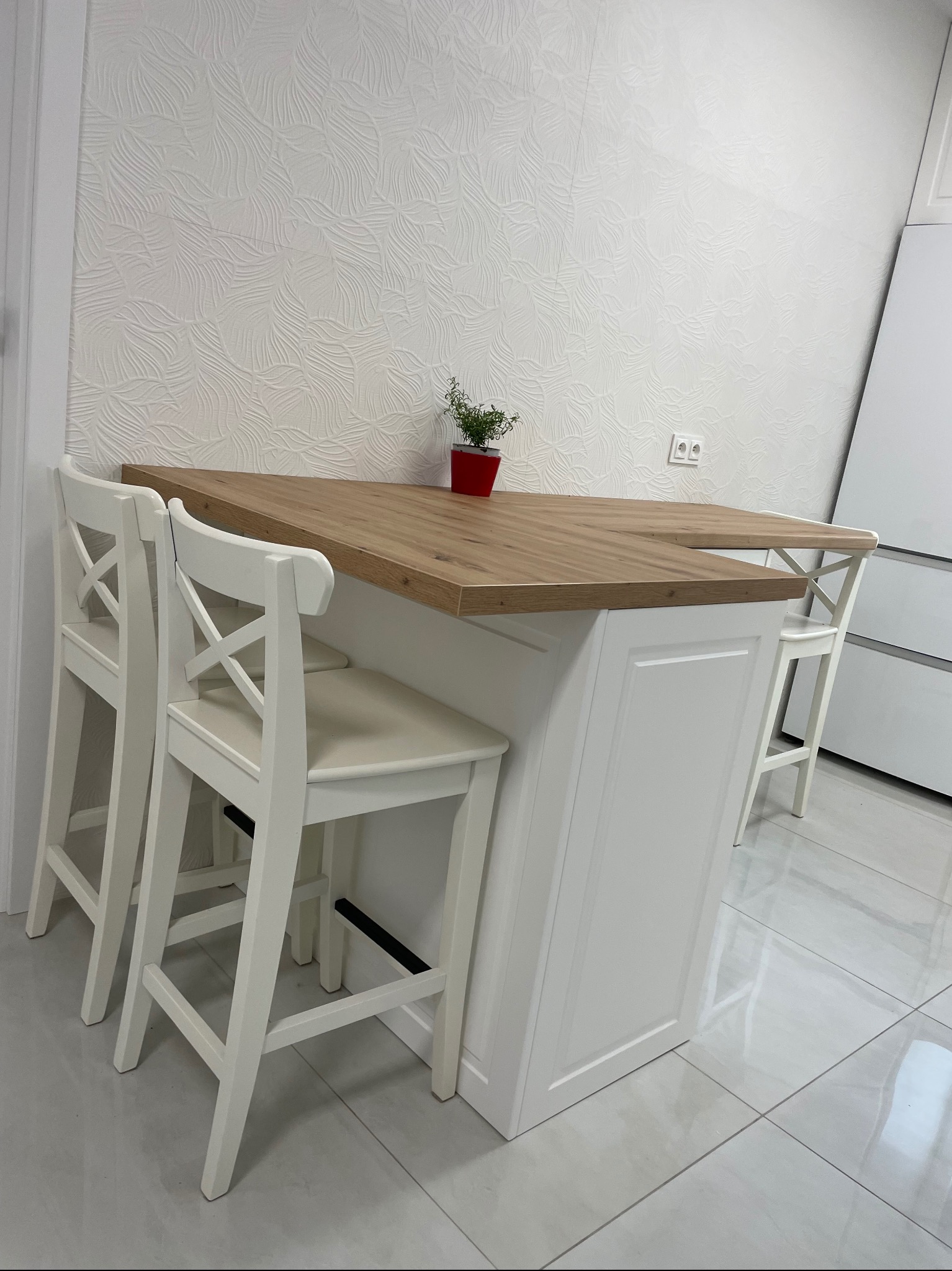 Nowoczesna wyspa kuchenna z drewnianym blatem i białymi krzesłami barowymi, uzupełniona czerwoną doniczką z ziołami na blacie, na tle białej ściany z teksturą.