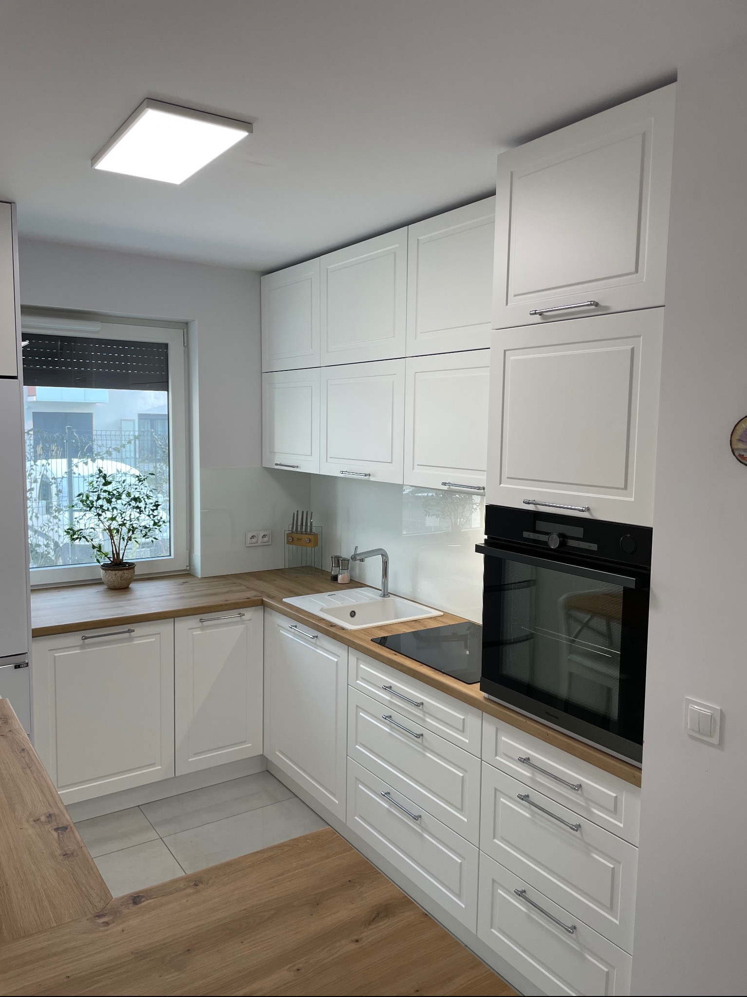 Nowoczesna, jasna kuchnia z białymi frontami szafek, drewnianym blatem i widokiem na zimowy krajobraz za oknem. Na blacie stoi doniczka z małym drzewkiem.