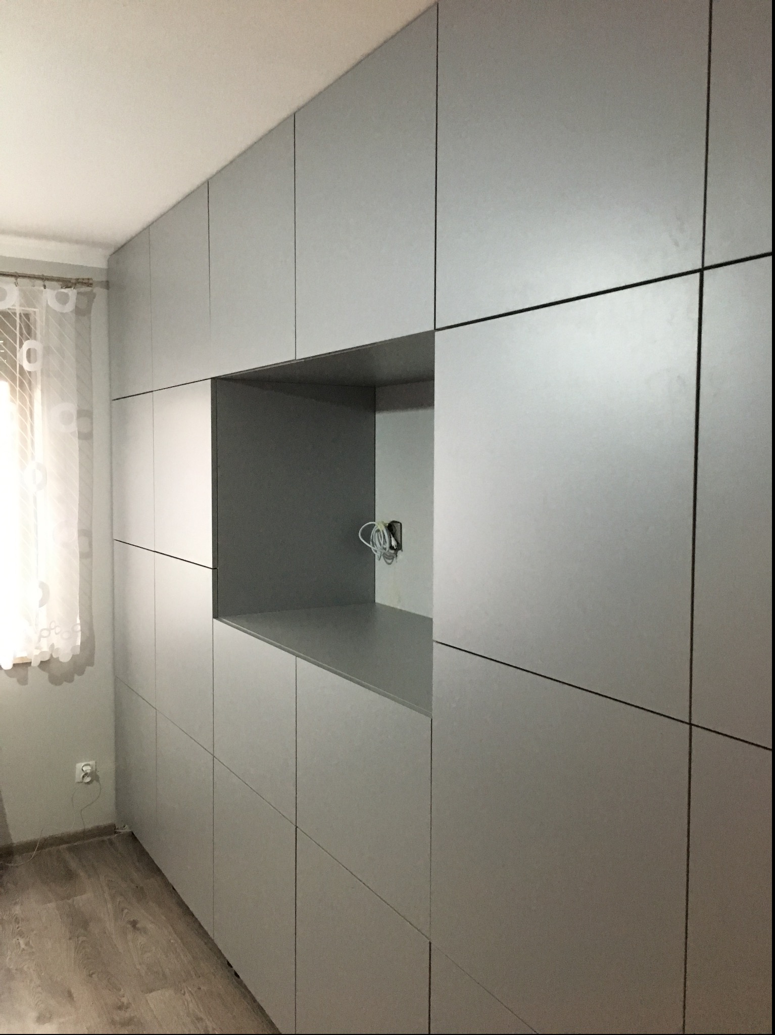 Szara zabudowa meblowa od podłogi do sufitu z otwartą wnęką na sprzęt RTV i wystającymi kablami, widoczna z boku przy ścianie z oknem z firanką.