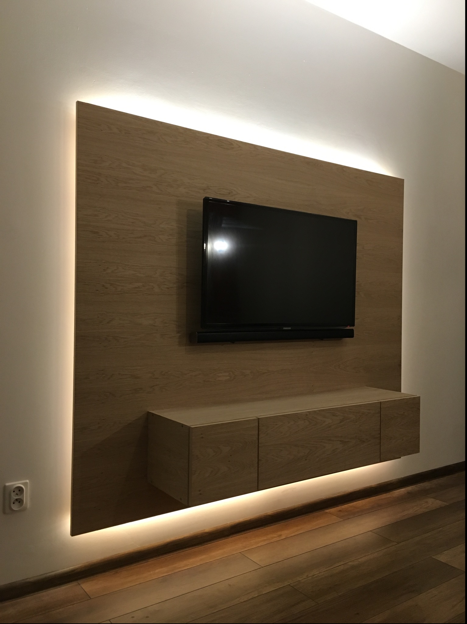 Drewniana zabudowa ściany z podświetleniem LED, podwieszana szafka RTV i telewizor na tle parkietu.