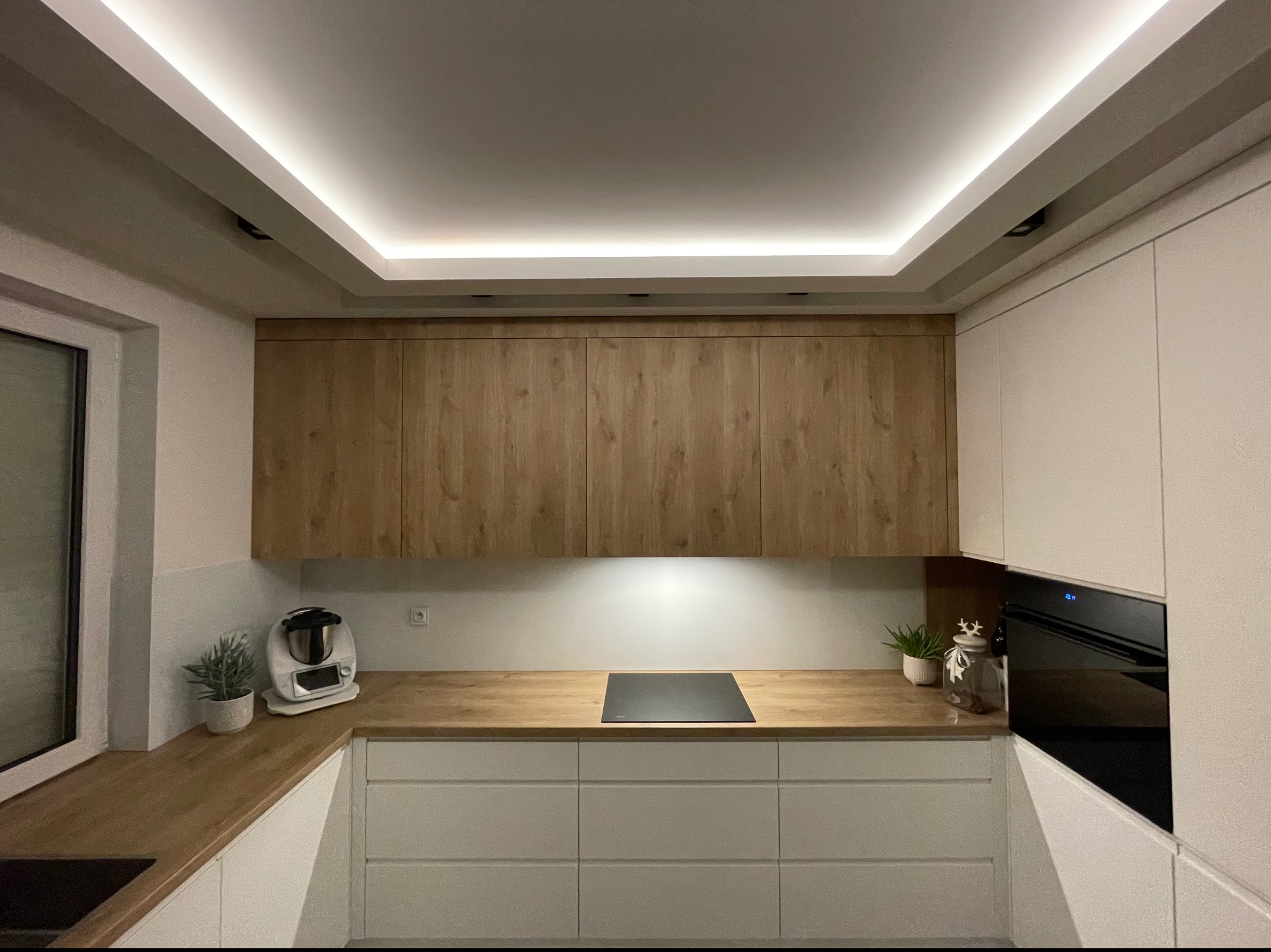 Nowoczesna kuchnia z białymi szafkami dolnymi i drewnianymi szafkami górnymi, blatem z drewna, oświetleniem podszafkowym i sufitowym LED, oraz sprzętem AGD.