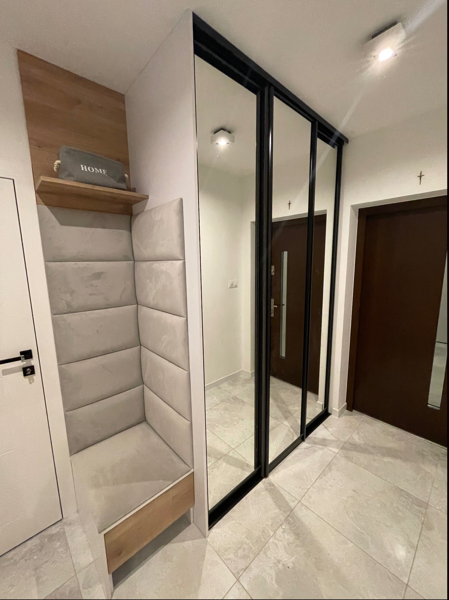 Nowoczesny przedpokój z zabudową siedziska z tapicerowanymi panelami, drewnianą półką i lustrzaną szafą przesuwną w czarnej ramie.