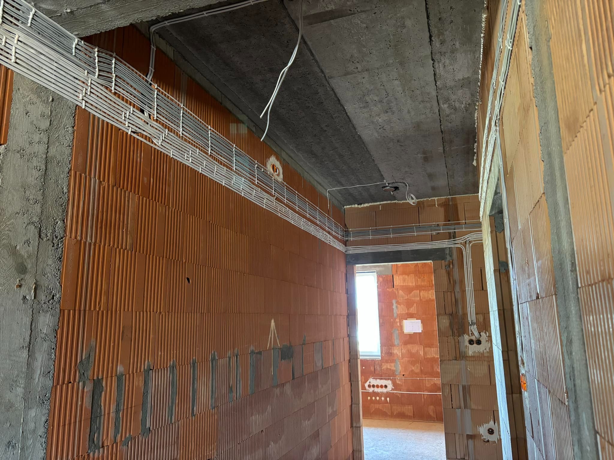 Instalacja elektryczna w budynku w trakcie budowy: kable prowadzone po ścianach z cegły, betonowy sufit. Widoczne puszki elektryczne i okno w tle.