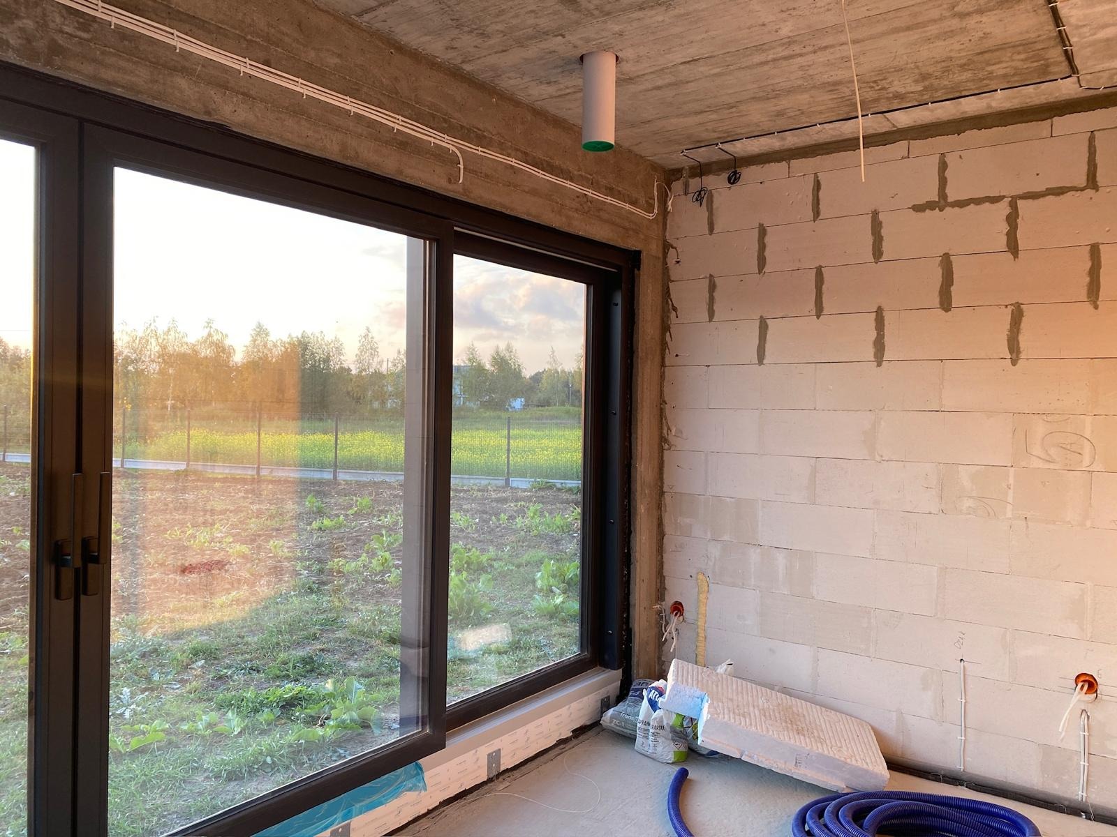 Surowy stan wnętrza z dużym oknem i widocznymi przewodami elektrycznymi pod sufitem. Ściany z betonu komórkowego, instalacja w trakcie realizacji.