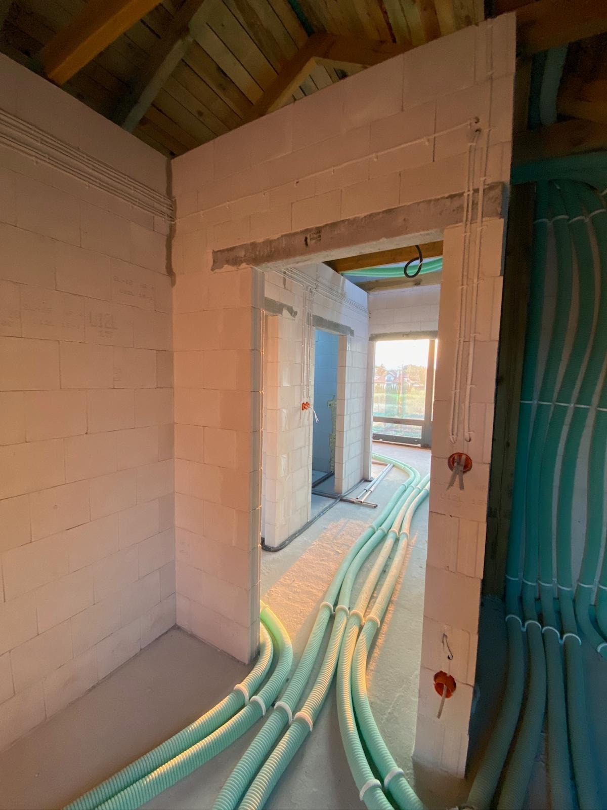 Wnętrze w trakcie budowy z widocznymi, ułożonymi rurami instalacyjnymi w podłodze i na ścianach. Surowy stan, ściany z białego betonu komórkowego, drewniany strop.