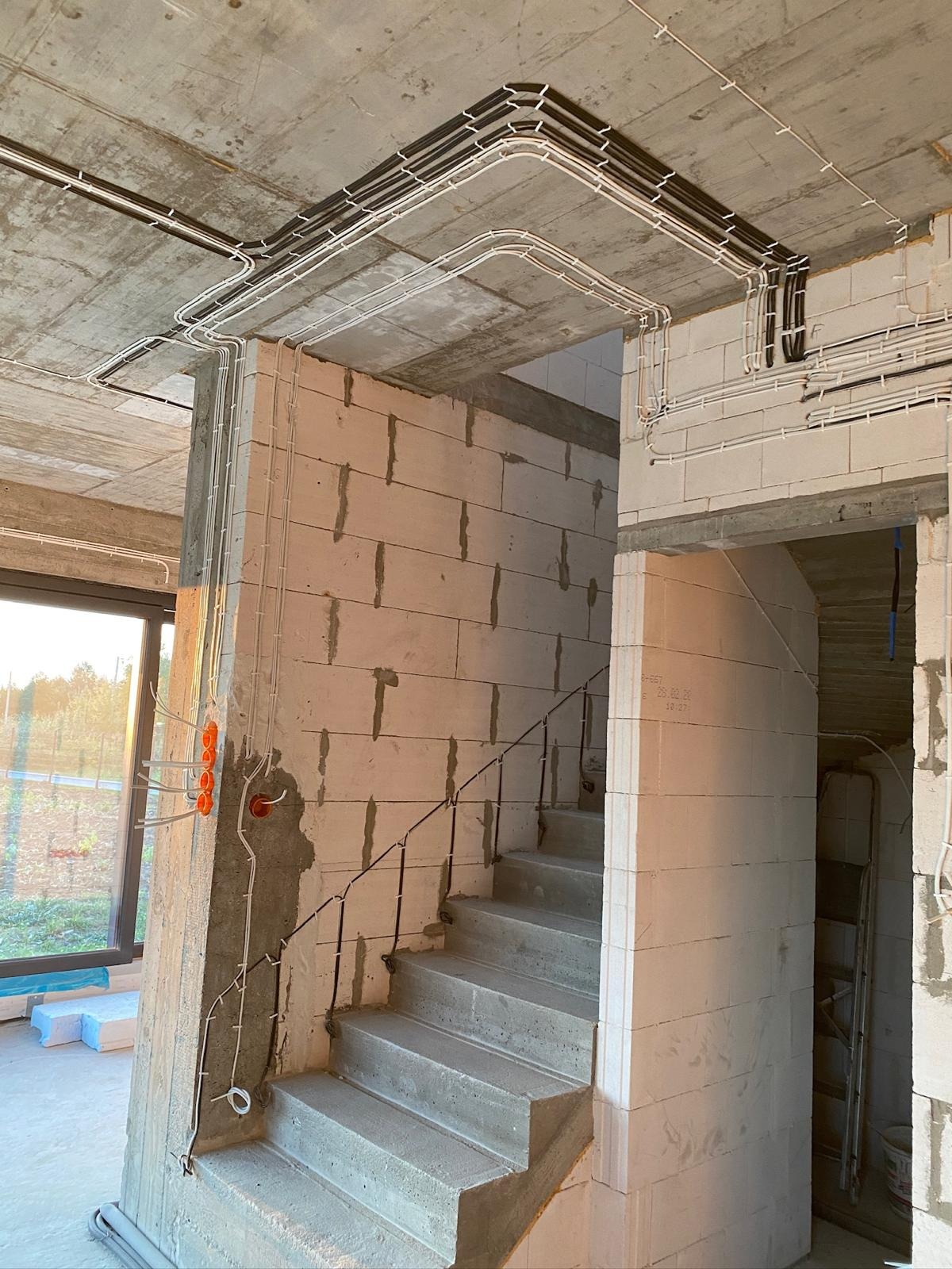 Surowy beton i instalacja elektryczna w budynku w trakcie budowy. Widoczne kable prowadzone po suficie i ścianach, puszki elektryczne, schody betonowe i okno z widokiem na zieleń.