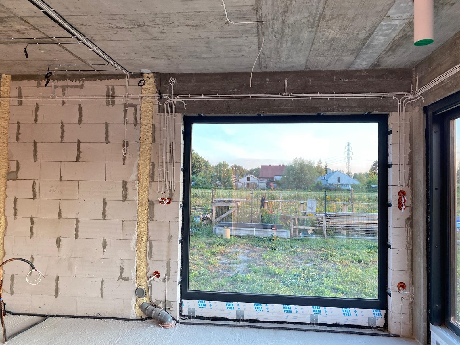 Surowy stan wnętrza z widoczną instalacją elektryczną w peszlach na ścianach z betonu komórkowego. Przewody prowadzone pod sufitem. Okno z widokiem na zieleń.