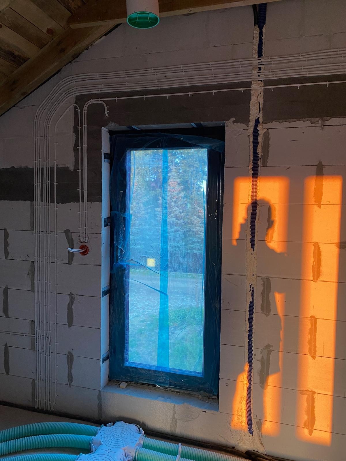 Instalacja elektryczna w surowym wnętrzu: białe kable ułożone wzdłuż ściany z betonu komórkowego, okno zabezpieczone folią, ciepłe promienie słońca padają na ścianę.