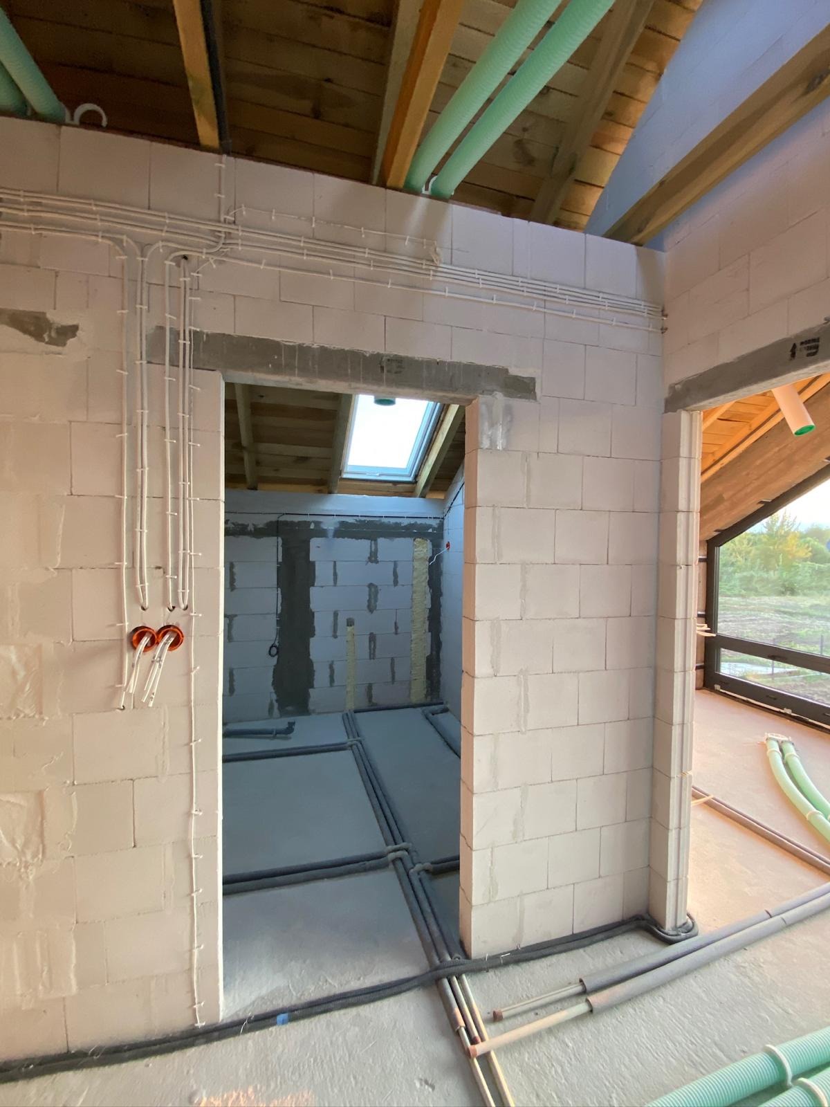 Surowy stan budynku z widoczną instalacją elektryczną i rurami ogrzewania podłogowego. Białe ściany z betonu komórkowego, widoczne kable i rury w podłodze.