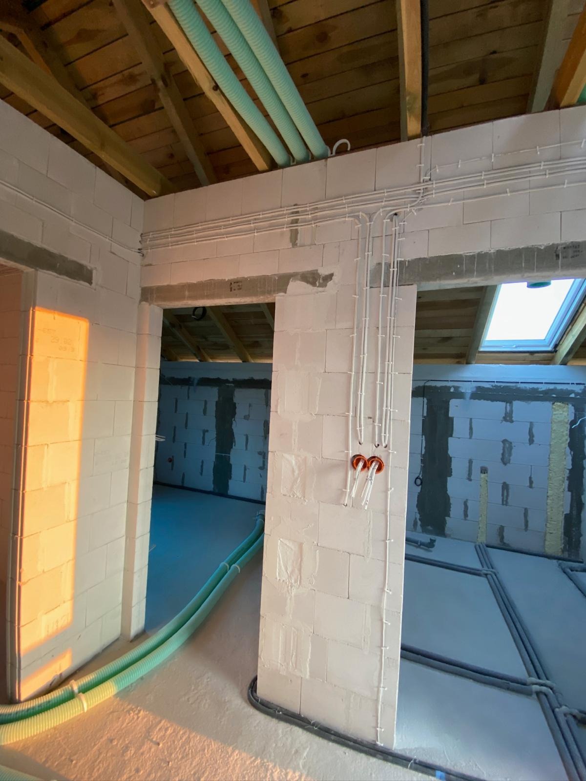 Wnętrze w trakcie budowy z widocznymi przewodami elektrycznymi w białych rurkach, poprowadzonymi po ścianach z białych bloczków i pod sufitem z drewnianych belek.