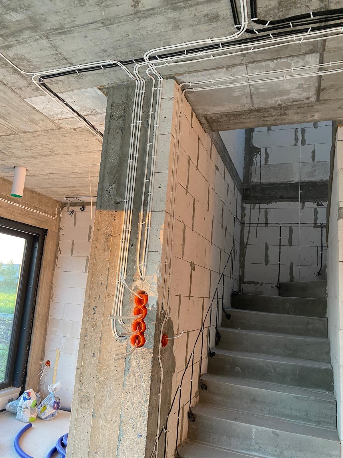 Surowy stan: kable elektryczne w białych rurkach prowadzone po suficie i ścianach w nowym budynku, pomarańczowe puszki podtynkowe, betonowe schody.